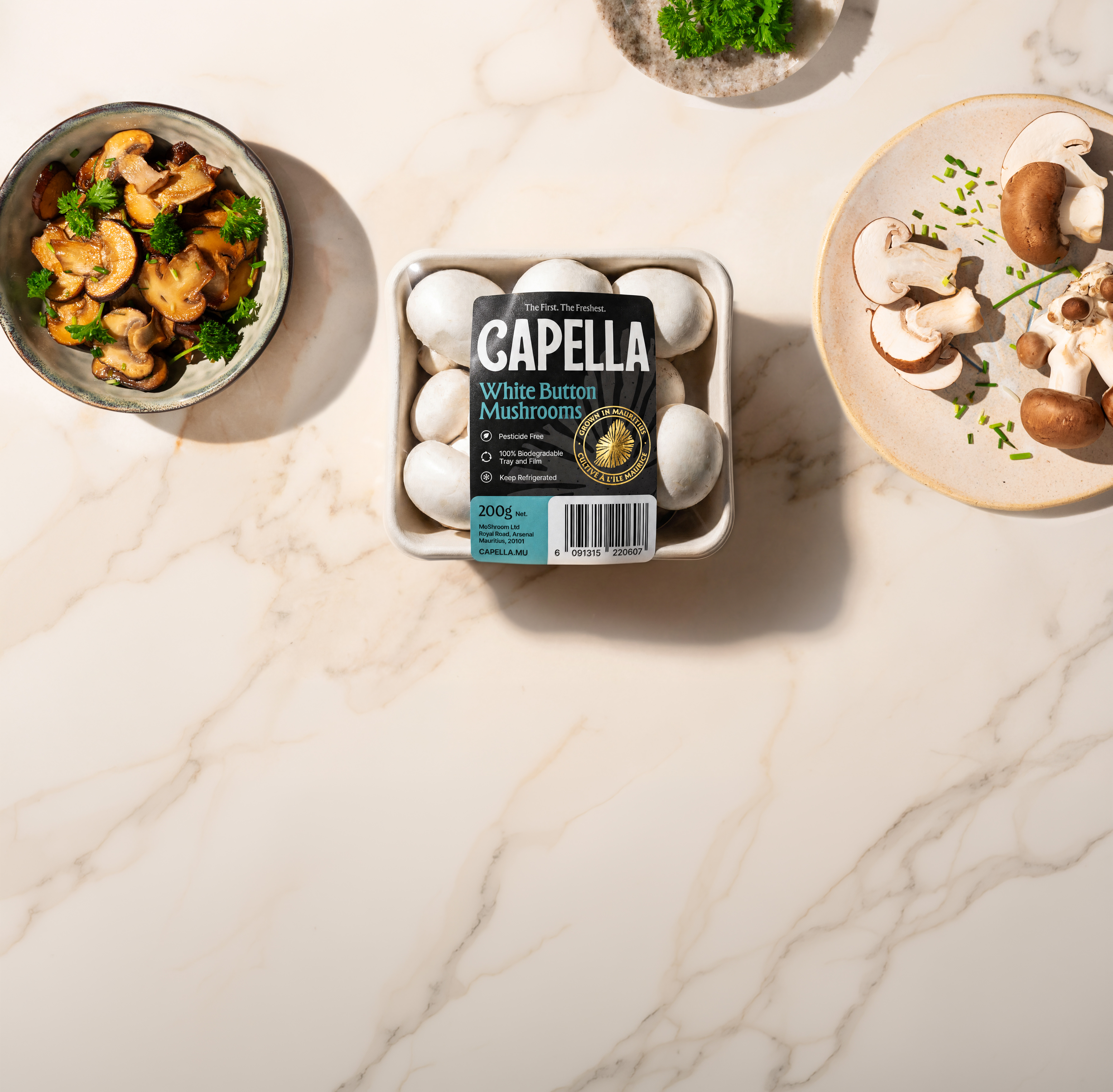Capella White Button Mushrooms in a tray on a marble surface, surrounded by cooked and sliced mushrooms